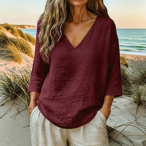 May include: A deep burgundy V-neck top with three-quarter length sleeves and a loose, relaxed fit, paired with light beige trousers. The top features a textured fabric finish and a simple, unadorned design with a straight hemline.