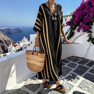 May include: A black kaftan dress featuring vertical gold stripes, a V-neckline, and wide sleeves. The garment is paired with a large, woven tan straw tote bag with horizontal stripes, a beaded necklace, and black flat sandals with wide straps.