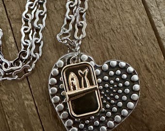 May include: A silver heart-shaped pendant necklace with a gold-toned charm of a sardine can with two fish inside. The pendant has a textured surface with small dots.
