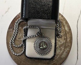 May include: A silver chain necklace with a round pendant featuring a silver and white design with the word "PARIS" in the center. The necklace is in a black box with a white interior.