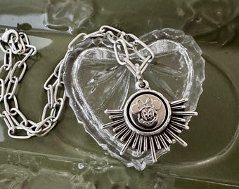 May include: Silver-toned necklace featuring a pendant with a Minnie Mouse design. The pendant has a sunburst design around the Minnie Mouse image. The necklace chain is a chunky link style. The necklace is resting on a clear heart-shaped object.