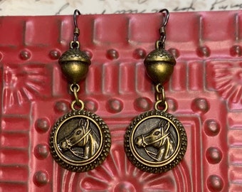 May include: A pair of gold-toned earrings with a horse head design. Each earring features a round pendant with a detailed horse head image and a smaller acorn-shaped bead above it.