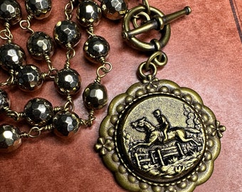 May include: A gold-toned necklace with a large pendant featuring a detailed image of a rider jumping a fence on horseback. The necklace is made of faceted beads and has a toggle clasp.