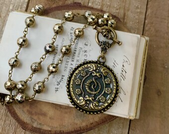 May include: A gold necklace with a large, round pendant featuring an ornate design and a green center. The pendant is attached to a chain made of small, faceted gold beads.