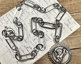 May include: A silver chain necklace with a large, round pendant featuring a cherub with wings. The pendant has a beaded border and the chain is made of interlocking oval links.