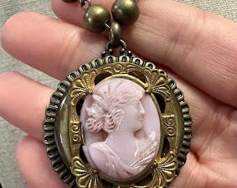 May include: A vintage-style necklace featuring a pink cameo pendant with a woman's profile. The cameo is set in a gold-toned, ornate frame and hangs from a beaded chain. The necklace is a classic piece of jewelry.
