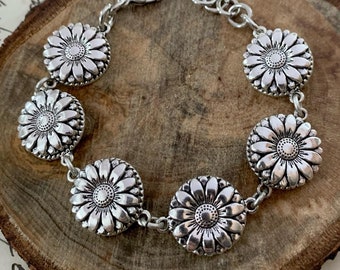 May include: A silver bracelet with a chain and six daisy-shaped charms. The bracelet is on a wooden surface.