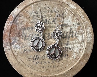 May include: Silver-toned dangle earrings featuring the Eiffel Tower within a circular frame. The earrings have ornate filigree accents and long ear wires. The background is a vintage metal container with faded text.