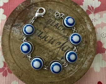 May include: A silver-toned bracelet featuring eight circular charms with a blue and white eye design. The charms are connected by small links. The bracelet is displayed on a rustic, circular metal surface with text. The background is a pink floral pattern.