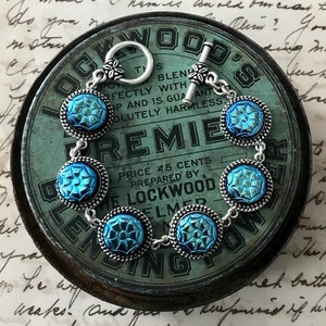 May include: A silver bracelet with a toggle clasp and six blue glass beads. The beads have a raised geometric pattern and are set in silver bezels. The bracelet is resting on a vintage tin with the text "Lockwood's Premier Blending Powder" printed on it.