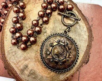 May include: A brown beaded necklace with a large, ornate, bronze pendant featuring a sunburst design and a woman's face. The pendant is attached to the necklace with a bronze toggle clasp.