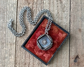 May include: A silver chain necklace with a square pendant featuring a silver flower design. The pendant is in a red velvet box.