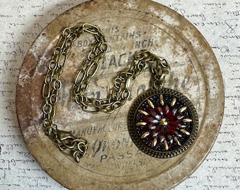 May include: A gold chain necklace with a round pendant featuring a red and gold floral design. The pendant is set in a brass frame with a textured finish.