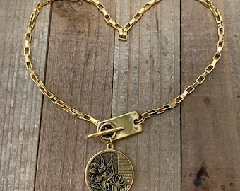 May include: A gold chain necklace with a toggle clasp and a round pendant featuring a floral design. The pendant is attached to the chain with a small, rectangular bar.