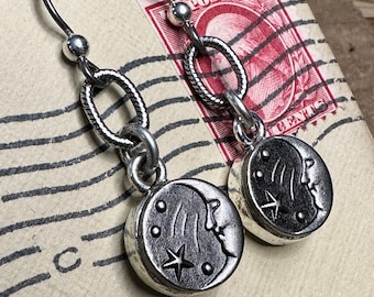 May include: A pair of silver earrings with a crescent moon design. Each earring features a small star and a crescent moon with a face. The earrings are hanging from a silver chain.