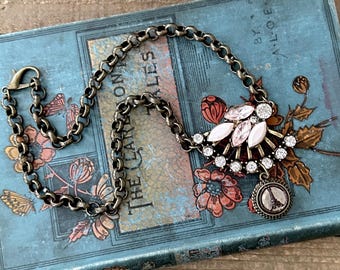 May include: Antique-style necklace with a bronze-toned chain and a decorative pendant. The pendant features a cluster of pink and clear crystals, with a small charm depicting the Eiffel Tower. The necklace rests on a vintage book with floral illustrations.