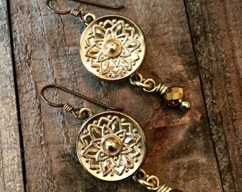 May include: Gold tone earrings with a round, ornate, floral design and a dangling brown crystal bead.