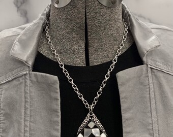 May include: A silver chain necklace with a teardrop-shaped pendant. The pendant is made of black and white patterned metal with a geometric design.