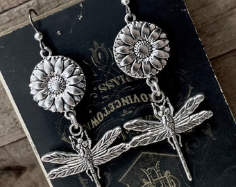 May include: Silver dangle earrings featuring a sunflower charm and a dragonfly charm. The sunflower charm is a round disc with a raised center and petals. The dragonfly charm is detailed with wings and a body.