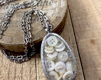May include: A silver-toned necklace with a pendant filled with white buttons. The pendant is teardrop-shaped with a decorative border. The necklace chain is also silver-toned, and the background is a rustic wooden surface.