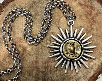 May include: Silver-tone necklace featuring a pendant with a sunburst design. The pendant has a circular center with an owl, moon, and star. The necklace is displayed on a wooden surface.