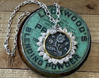 May include: Silver-toned necklace with a sunflower pendant. The pendant features a floral design with a gold-colored rim and a small ladybug charm. The necklace rests on a vintage green tin with text.