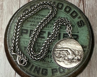 May include: A silver chain necklace with a silver pendant featuring a woman swimming in the ocean waves. The necklace is laying on top of a vintage green tin with the text "WOOD'S PREMIER POLISHING POWDER" printed on it.