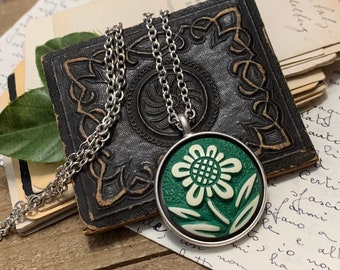 May include: A silver chain necklace with a green and white flower pendant. The pendant is a round, ceramic button with a raised flower design.