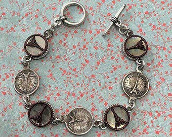 May include: Silver-toned bracelet featuring circular charms with the Eiffel Tower. The charms alternate between a detailed tower image and a coin-like design with the words "LA TOUR EIFFEL". The bracelet has a toggle clasp.