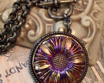 May include: A vintage-style pendant necklace featuring a large, round, iridescent glass pendant with a floral design in shades of gold and purple. The pendant is set in a bronze-toned metal frame and hangs from a matching chain with a toggle clasp.