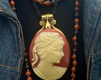 May include: A gold-toned pendant necklace featuring a cameo of a woman's profile in cream against a red background. The pendant is suspended from a black chain, complemented by strands of orange and black beaded necklaces. The jewelry is set against a dark background.