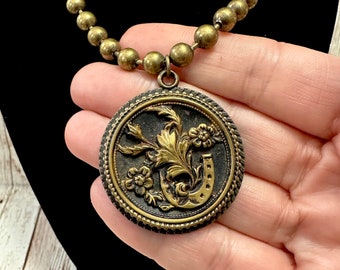 May include: A gold necklace with a large round pendant featuring a horseshoe and floral design. The pendant is made of brass and has a dark brown finish.