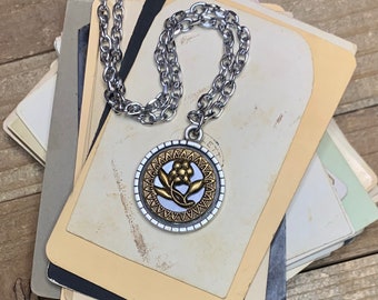 May include: A silver chain necklace with a gold pendant featuring a stylized flower design. The pendant has a white center and a gold rim with a decorative pattern.