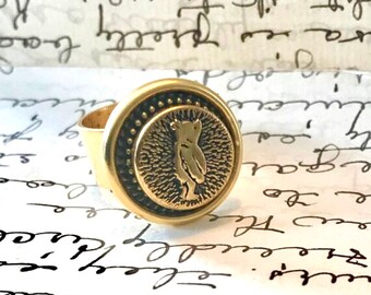 May include: A gold-toned ring featuring a detailed owl design. The ring's face has a circular shape with an embossed owl in the center, surrounded by a textured border. The band is a simple, polished gold.
