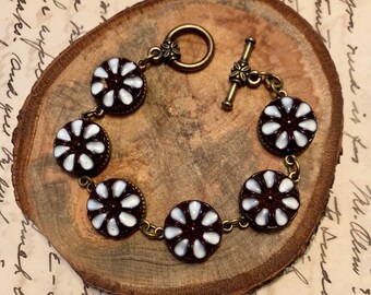May include: A brown and white floral bracelet with a brass clasp and chain. The bracelet has six round floral beads with white centers and brown edges.