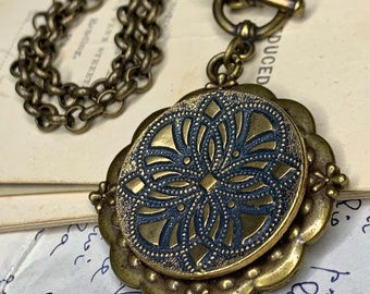 May include: A brass necklace with a round pendant featuring a black and gold geometric design. The pendant has a scalloped edge and is attached to a chain with a toggle clasp.