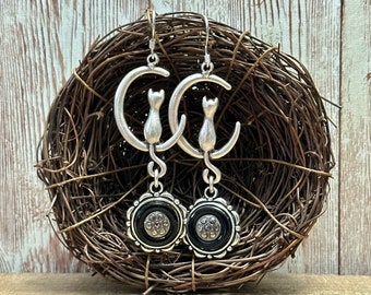 May include: Silver dangle earrings with a crescent moon design and a black cat silhouette. The earrings feature a black and silver button-like charm.