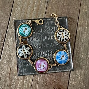 May include: A gold chain bracelet with six colorful ceramic buttons. The buttons have different patterns, including a flower, a geometric design, and a star.