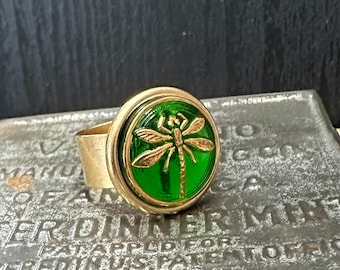 May include: A gold ring with a green glass cabochon featuring a gold dragonfly design.
