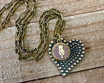 May include: A gold necklace with a heart-shaped pendant. The pendant has a textured surface and a small round charm with a bear silhouette in the center.
