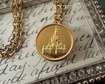 May include: Gold-toned necklace featuring a round pendant with a castle design. The pendant hangs from a chain with a textured link pattern. The necklace is displayed on a background with script writing and a decorative frame.