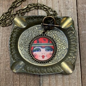 May include: A gold chain necklace with a round pendant featuring a vintage illustration of a woman with red hair and green eyes. The pendant is surrounded by a beaded border.