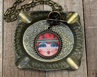 May include: A gold chain necklace with a round pendant featuring a vintage illustration of a woman with red hair and green eyes. The pendant is surrounded by a beaded border.
