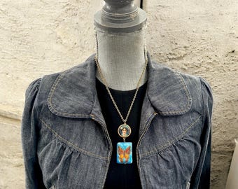 May include: A gold-toned necklace with two pendants. The top pendant is round with a portrait, and the bottom pendant is rectangular with a butterfly design. The necklace is displayed on a mannequin wearing a dark denim jacket.