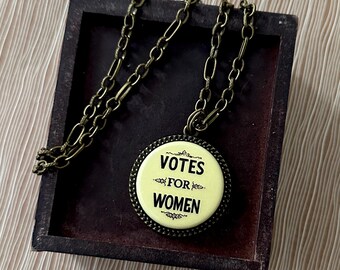 May include: A vintage-style necklace with a round pendant that reads "VOTES FOR WOMEN" in black text on a pale yellow background. The pendant is framed with a decorative border and hangs from a bronze-colored chain.
