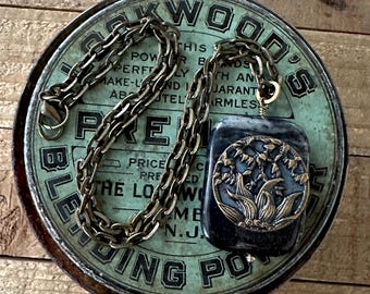 May include: A vintage-style necklace with a dark blue rectangular pendant and a bronze-colored floral design. The pendant hangs from a bronze chain. The necklace rests on a distressed, round, teal-colored metal container.