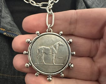 May include: A silver-toned pendant necklace featuring a circular coin-style charm with a horse relief design. The charm is framed by small metallic spheres along the edge and hangs from a thick, interlocking silver-toned chain link necklace.
