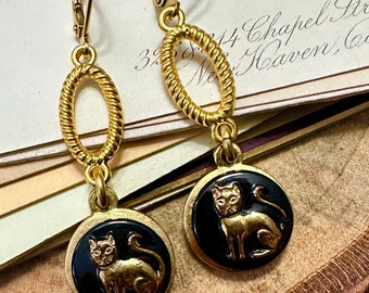 May include: Gold-tone leverback earrings. Each earring features a gold oval link and a black circular pendant with a gold cat design. The earrings are displayed on a wooden surface with vintage paper in the background.
