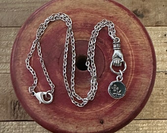 May include: A silver chain necklace with a silver hand charm and a silver charm with a floral design.