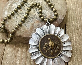 May include: A silver and gold necklace with a large, ornate flower pendant featuring a bird on a branch. The necklace is made of brass beads.